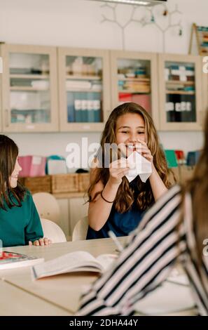 Smiling female student holding paper craft while sitting by table in classroom Stock Photo