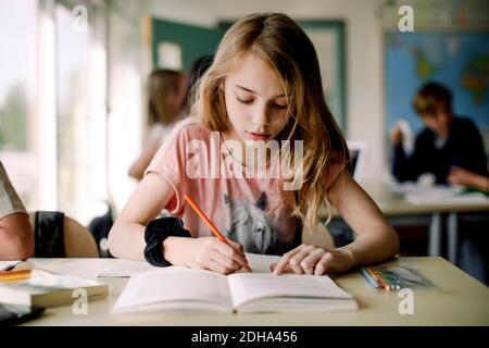 Female student writing in book while sitting at table in classroom Stock Photo