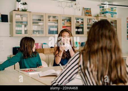Smiling female student holding paper craft while sitting by friend in classroom Stock Photo