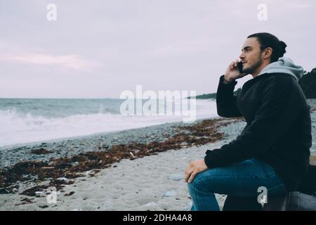 Side view of young man using mobile phone while sitting at beach Stock Photo