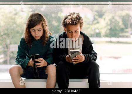 Male and female students using mobile phone while sitting against glass window in school Stock Photo