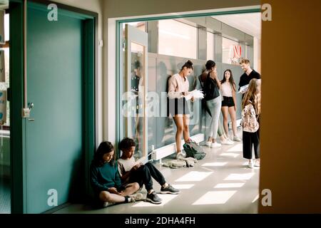 Girl and boy sitting in doorway while professor talking to students in school corridor