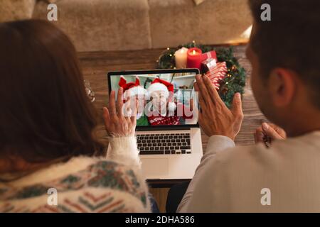 Rear view of couple waving while having a videocall with senior couple in santa hats smiling on lapt Stock Photo