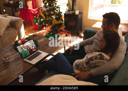 Couple sitting on couch having a videocall with senior couple in santa hats smiling on laptop at hom Stock Photo