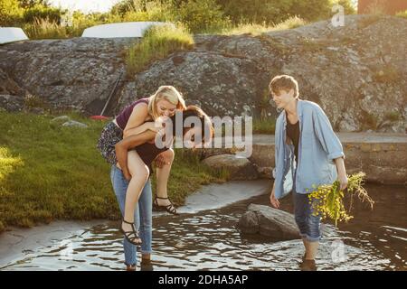 Man looking at young woman giving piggyback ride to female friend while walking in water Stock Photo