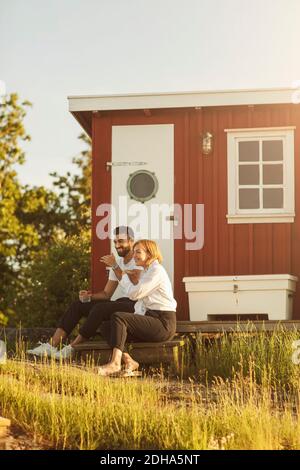 Happy couple sitting at log cabin on sunny day Stock Photo