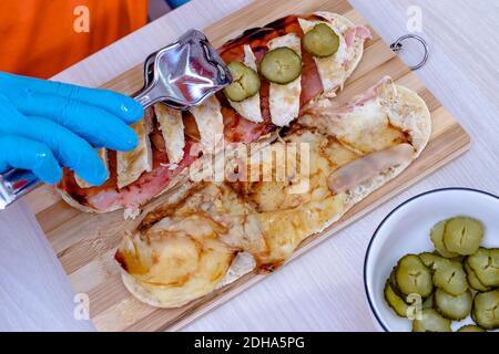 Chef preparing tasty fresh baguette submarine sandwich. Close up of hands in gloves holding tongs and arranging slices of pickle over the meat. Stock Photo