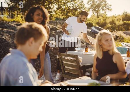Young multi-ethnic friends sitting while man setting table at harbor on sunny day Stock Photo