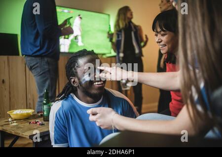 Young woman applying face paint on friend's cheek during soccer match Stock Photo