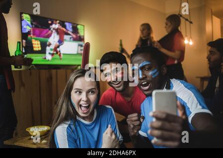 Fans taking selfie with mobile phone while friends watching soccer match on TV Stock Photo