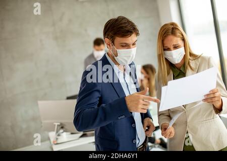 Young man and woman with protective facial masks discussing with paper in hands indoors in the office with young people works behind them Stock Photo