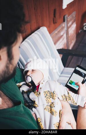 Father with sleeping baby boy using mobile phone while sitting on chair Stock Photo