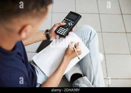 Young man using calculator and writing on paper while sitting with crossed legs at laundromat Stock Photo