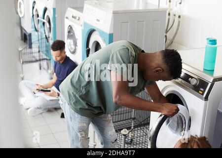 Man using washing machine while university student studying at laundromat Stock Photo