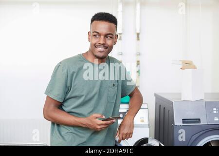 Smiling man holding mobile phone while standing at laundromat Stock Photo