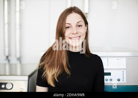 Portrait of smiling young woman with long brown hair standing at laundromat Stock Photo
