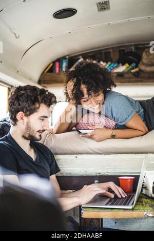 Smiling young woman lying on bed while looking at man using laptop in motor home Stock Photo
