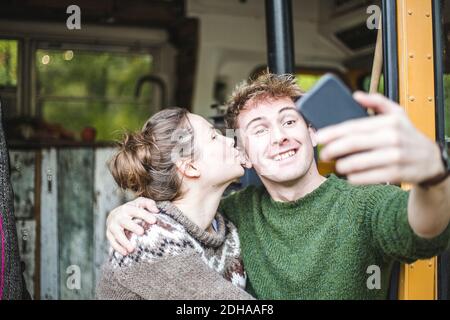 Smiling man taking selfie from mobile phone while woman kissing him during camping Stock Photo