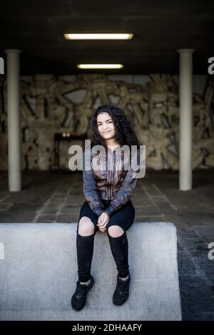 Full length portrait of teenage girl sitting on concrete at parking garage Stock Photo