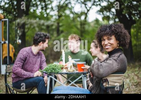 Portrait of smiling Afro woman sitting with friends at table during camping in forest Stock Photo