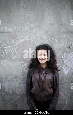 Portrait of smiling teenage girl standing against chalk drawing on gray wall in city Stock Photo