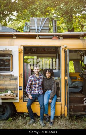 Portrait of smiling young multi-ethnic couple sitting at camper vehicle doorway during camping in forest Stock Photo