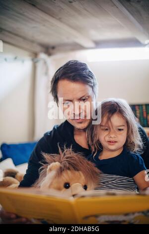 Father reading book with daughter while sitting in bedroom Stock Photo