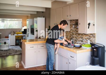 Father washing kitchen utensil while daughter sitting on kitchen counter Stock Photo