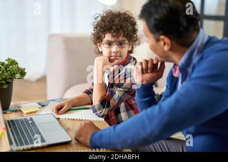 Cute latin school boy looking thoughtful, talking to his father, discussing his homework while sitting at the table at home. Remote studying, family, fatherhood concept. Selective focus Stock Photo