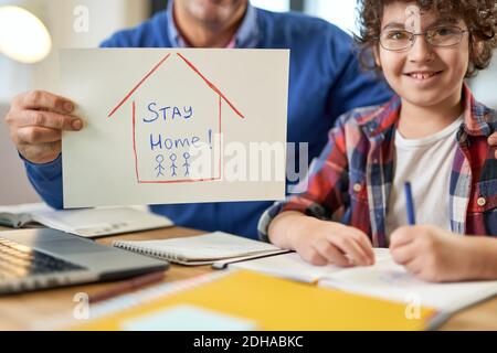 Happy latin boy sitting at the desk together with his father during remote learning at home. Dad holding Stay home drawing. Online education, homeschooling concept Stock Photo