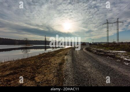 Rural landscape with dark brown gravel road between lake and a field with high-voltage power lines. Cloudy winter evening with blue sky, sun hiding. Stock Photo