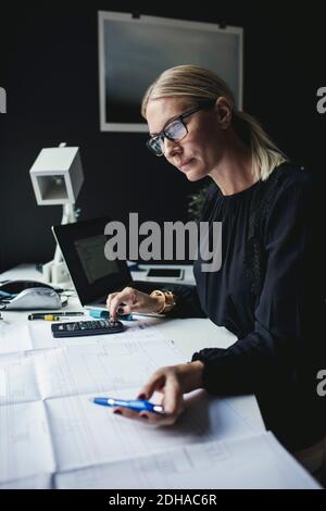 Businesswoman looking at blueprint while using calculator on desk Stock Photo