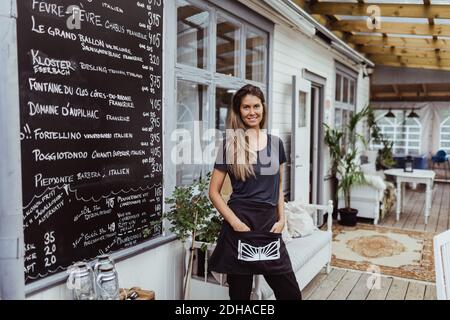 Portrait of smiling female owner with hands in pockets standing in restaurant Stock Photo