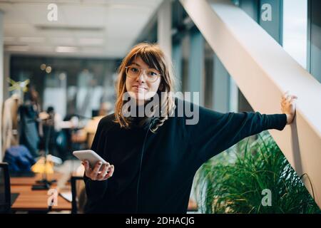 Portrait of confident businesswoman standing with smart phone in office Stock Photo