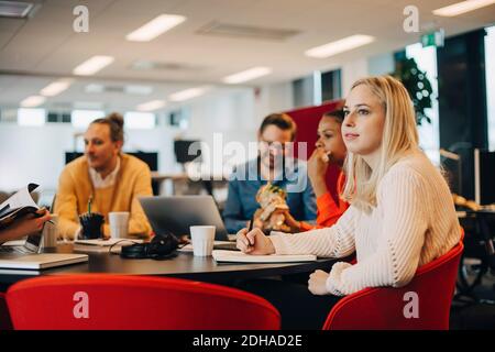 Thoughtful young businesswoman looking away while sitting with colleagues at conference table in office Stock Photo