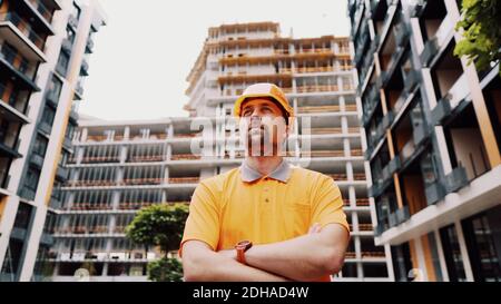 Young caucasian constructor with arms crossed in safety helmet and orange reflective uniform posing with proud expression at con Stock Photo