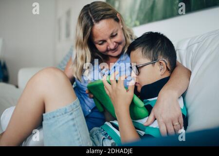 Smiling mother looking at disabled son using digital tablet while sitting on sofa Stock Photo
