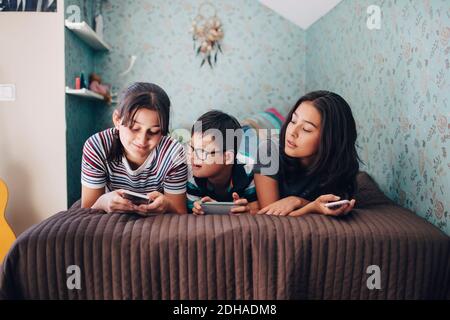 Siblings using wireless technologies while lying on bed by wall at home Stock Photo