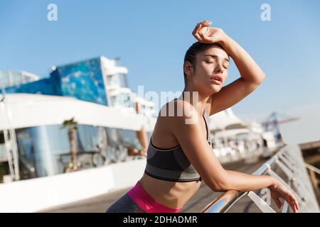 Close-up of attractive young fitness woman leaning on handrail, wiping sweat off forehead after workout, finish jogging on the s Stock Photo