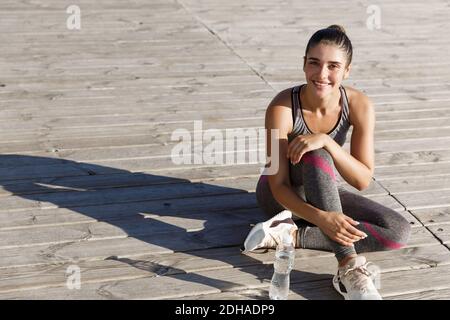Outdoor shot of happy sportswoman having break after workout or jogging, sitting in sports clothing on wooden pier and smiling a Stock Photo