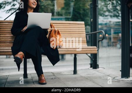 Low section of smiling mid adult businesswoman sitting with laptop at bus shelter Stock Photo