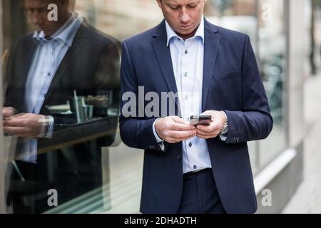 Contemplating mature businessman using smart phone while standing by glass window Stock Photo