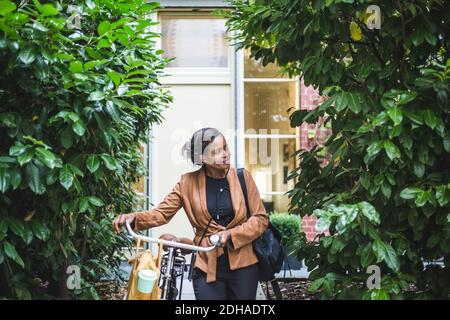 Smiling female architect looking away while standing with bicycle against house Stock Photo