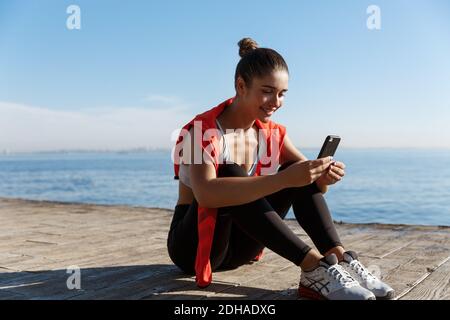 Outdoor shot of happy fitness woman sitting on wooden pier and smiling, using smartphone, having break after workout near the se Stock Photo