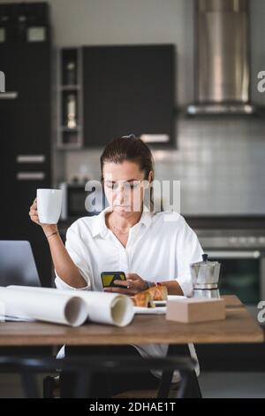 Female freelancer using phone while sitting with coffee cup at home Stock Photo