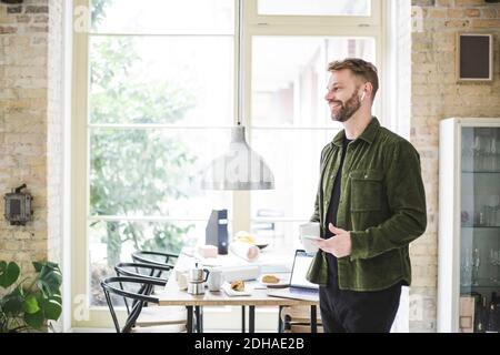 Smiling entrepreneur looking away while holding phone and cup at home Stock Photo