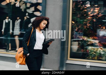 Mid adult businesswoman walking while using digital tablet with hand in pocket against building in city Stock Photo