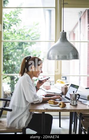 Smiling female freelancer using laptop while talking through smart phone at home office Stock Photo