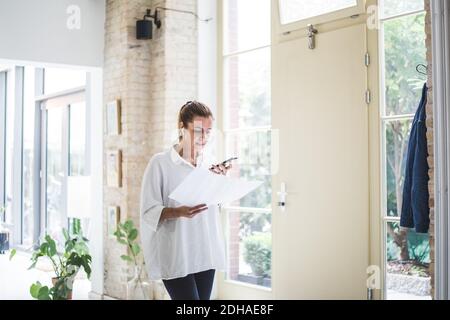 Female designer talking through smart phone while looking at document in home office Stock Photo