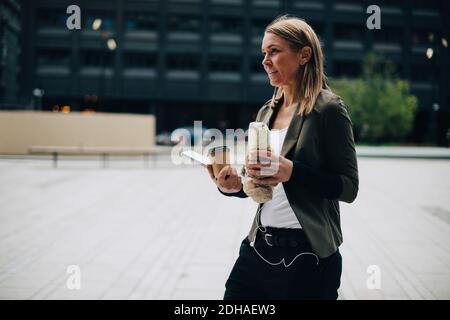 Mature businesswoman carrying food and drink with laptop while walking on footpath in city Stock Photo